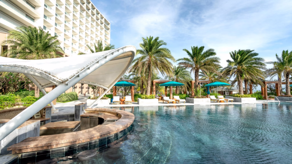 a luxury hotel and resort exterior with a wide pool, lounge chairs, and trees